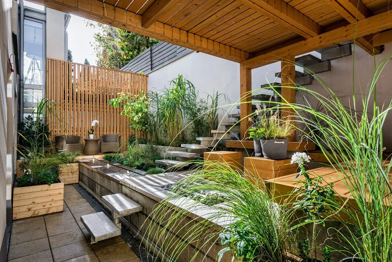 Small outdoor patio with lush green plants, wooden deck and stairs, sitting area with wicker chairs, and a privacy wooden fence.