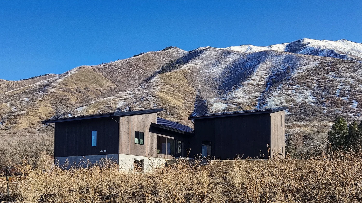 Modern black and wood-paneled house in a mountainous landscape with snow patches and sparse vegetation, under a clear blue sky.