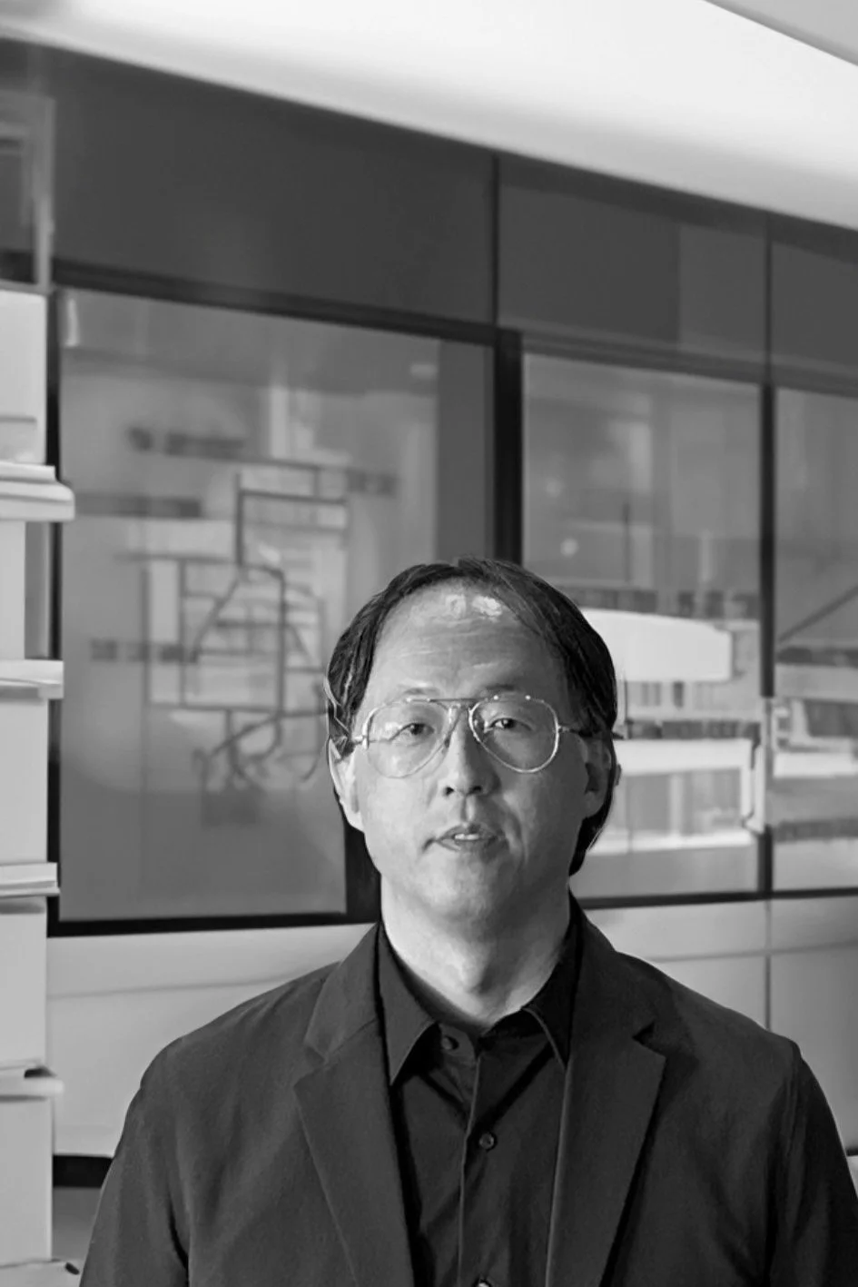 Black and white photo of a man with glasses in a suit standing in an indoor space with glass walls and shelves.