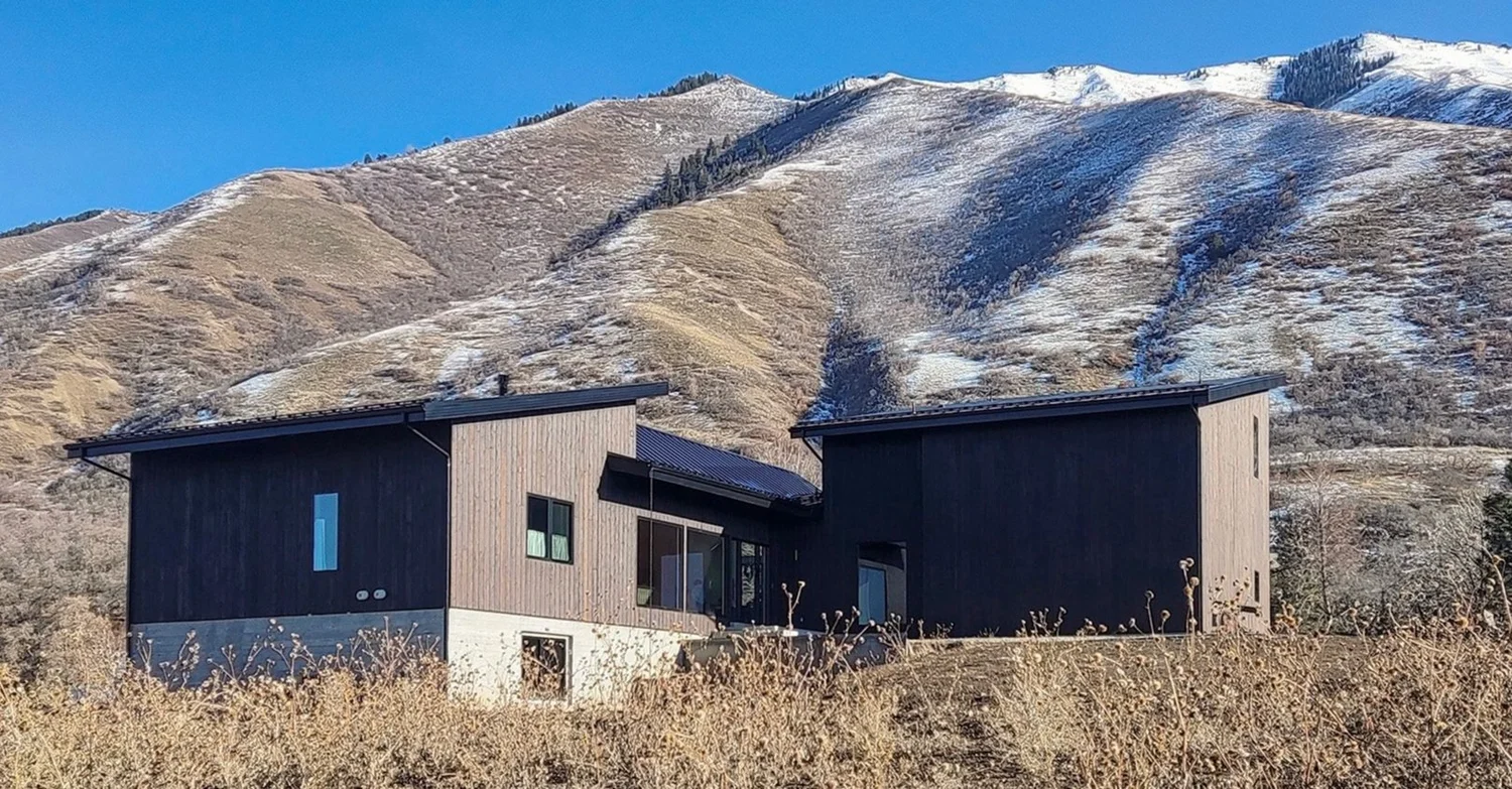 Modern black and gray house with large windows in a mountainous area with brown and snow-covered slopes.
