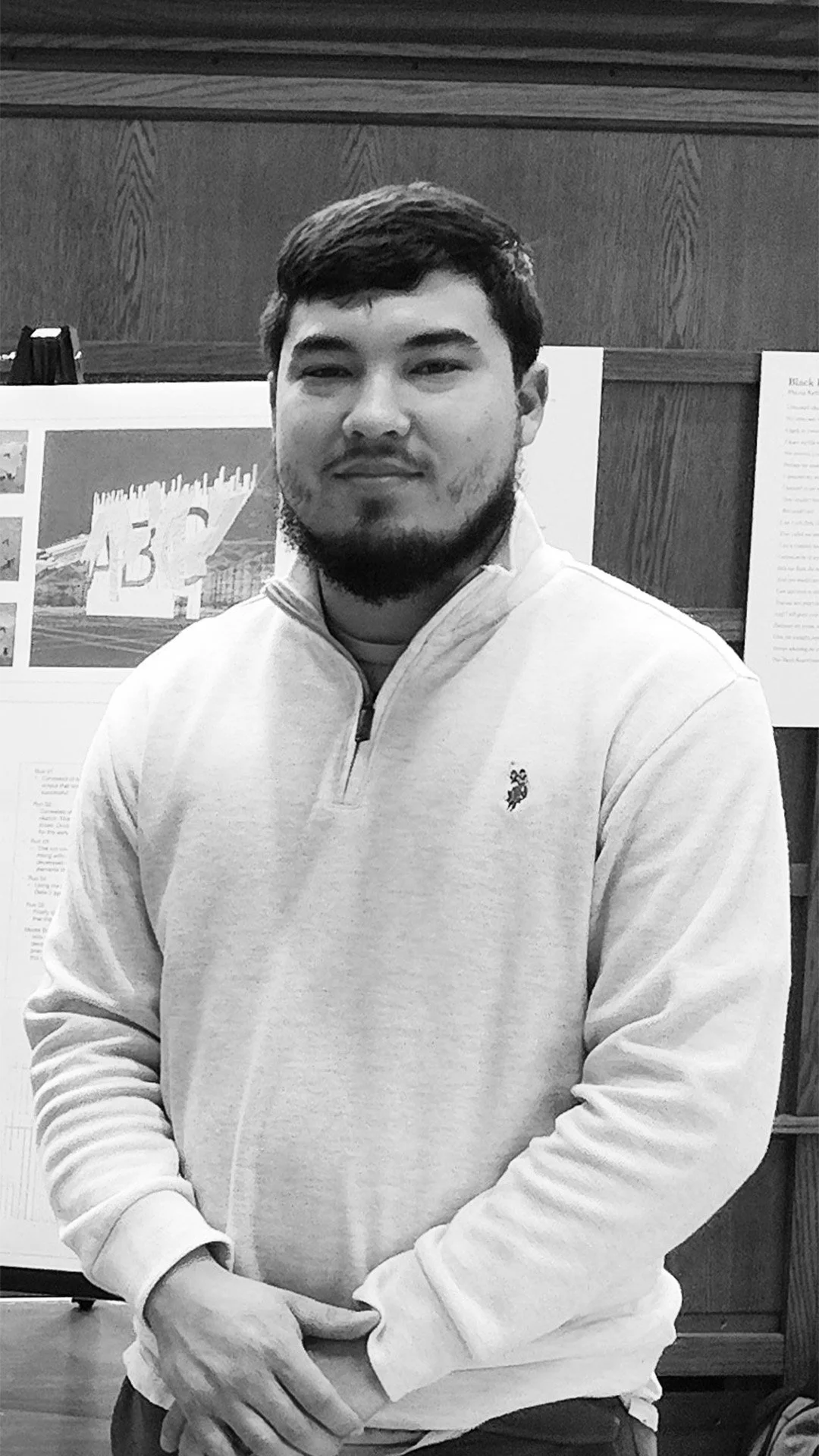 A man with dark hair and a beard, wearing a light-colored zip-up sweatshirt, sitting in front of a display board with information and images.