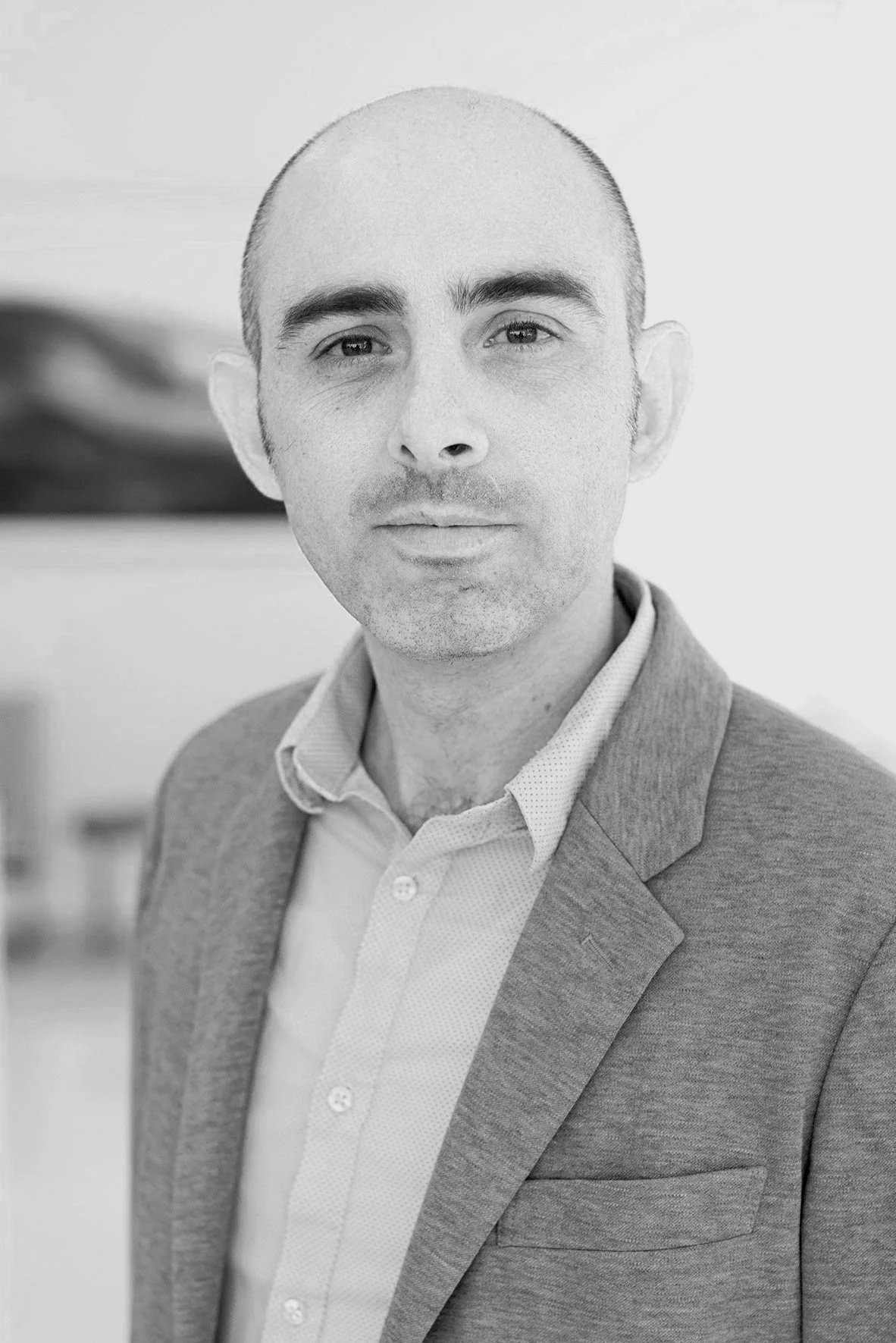 A black and white portrait of a man with a shaved head, wearing a suit jacket and collared shirt, with a serious expression, standing indoors.