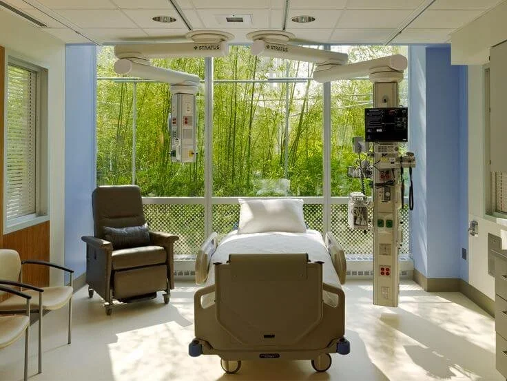 Hospital room with a bed, medical equipment, a brown recliner chair, and large windows showing green trees outside.